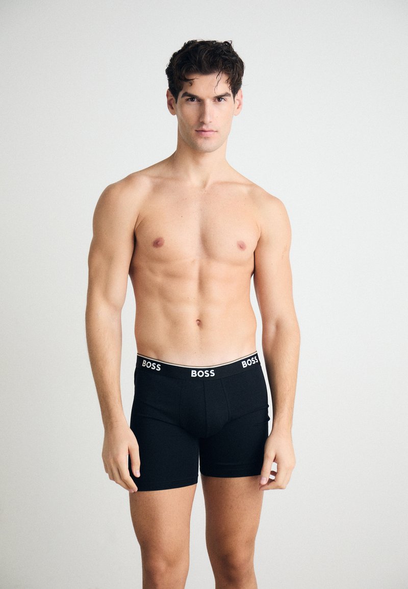 Schwarze Boxer-Shorts mit einem weißen "BOSS"-Logo am Bund. Das Material wirkt dehnbar; das Modell steht mit einem neutralen Ausdruck.