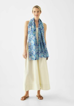 Vrouw die een mouwloze crèmekleurige maxi-jurk, bruine sandalen en een lange blauwe sjaal met paisley-patroon draagt, staand tegen een witte achtergrond.