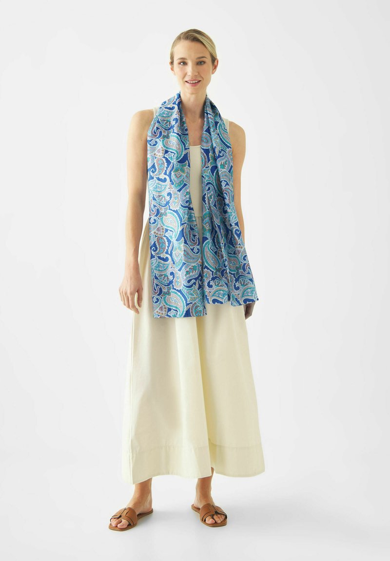 Vrouw die een mouwloze crèmekleurige maxi-jurk, bruine sandalen en een lange blauwe sjaal met paisley-patroon draagt, staand tegen een witte achtergrond.