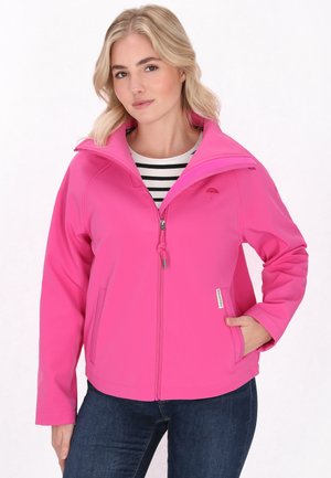 Una donna con capelli biondi mossi indossa una giacca rosa brillante sopra una camicia a righe, posando con una mano in tasca su uno sfondo chiaro.