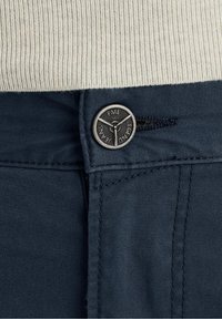 Pantaloni blu navy con una superficie liscia, dotati di un bottone metallico rotondo con il nome del marchio inciso e chiusura con zip in vita.