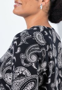 Schwarzes Oberteil aus Stoff mit einem weißen Paisleymuster, runder Ausschnitt und lockerem Schnitt. Auffällige silberne Ohrringe setzen Akzente.