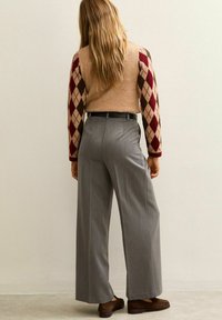 Maglione marrone chiaro con maniche a rombi rosse e crema, abbinato a pantaloni grigi a vita alta e larghi e scarpe marroni. Accessori minimi.