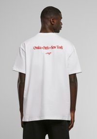White cotton t-shirt featuring red text on the back reading "Osaka o Paris o New York" with a small graphic element below.