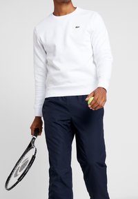 Vit sweatshirts med rund halsringning och ribbade ärmslut, i kombination med marinblå byxor, håller en tennisboll och racket. Grön logotyp som accent.