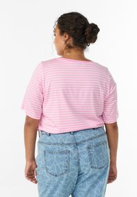 Kvinde med mørkt, krøllet hår i en knold iført sølvfarvede hoops, en lyserød og hvid stribet skjorte og lyseblå mønstrede jeans, som vender væk.