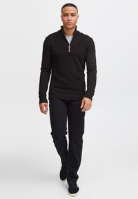 Black knit pullover with a quarter zip and ribbed cuffs, paired with dark jeans and black sneakers featuring white stripes on the side.