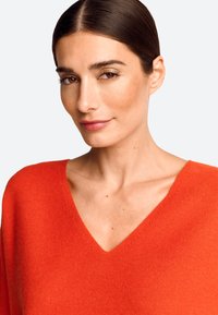 V-hals trui in fel oranje, gemaakt van zachte stof. Heeft een losse pasvorm met zichtbare textuur en een gladde halslijn. Geen patronen of accenten.