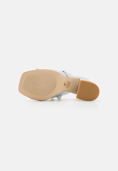 Sandale argentée à talons avec une semelle en caoutchouc beige texturé et une tige de couleur claire. Présente des larges sangles et des accents décoratifs en métal.
