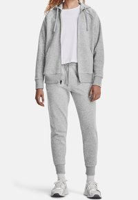 Sudadera gris con bolsillos y cordones, combinada con pantalones de chándal a juego con puños acanalados. Llevada con zapatillas blancas y grises.