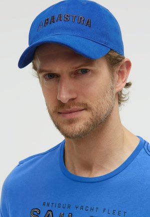 Man with light beard wearing a blue cap with "GAASTRA" logo and a matching blue t-shirt with "Antigua Yacht Fleet" text.