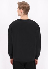 Fekete pulóver hosszú ujjakkal, kerek nyakkivágással és bordás mandzsettákkal. Simaság jellemzi, látható minták nélkül, hátulról nézve.