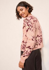 Rozā džemperis ar ziedu rakstu tumšākās toņos. Mīksta tekstūra, ribota aproce un īsa piegriezuma dizains. Valkāts kopā ar tumšām, piegulošām biksēm.