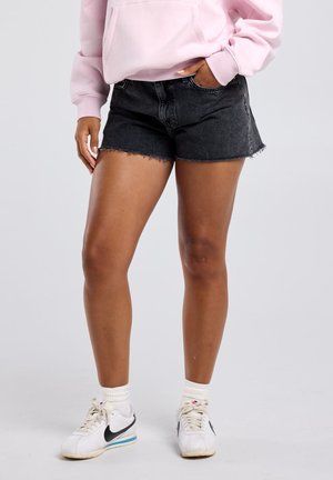 Persoon die vervaagde zwarte denimshorts draagt, lichtroze hoodie, witte Nike sneakers met sokken, staand met één hand in de zak tegen een effen achtergrond.
