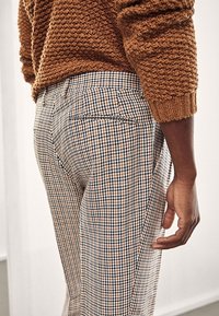 Personne portant un pull marron texturé et un pantalon taille haute à motif pied-de-poule, debout avec la main droite détendue le long du corps.