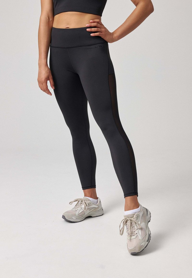 Leggings noirs avec une taille haute, ornés de panneaux latéraux transparents. Associés à des chaussures de sport gris clair. Texture lisse.