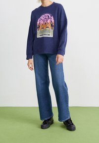 Marineblaues Sweatshirt mit grafischem Druck, der einen Sonnenuntergang und Palmen zeigt, kombiniert mit hellblauen weiten Jeans und schwarzen Schnürschuhen.