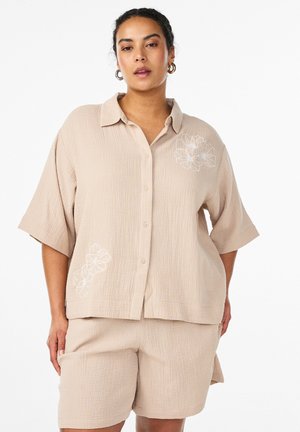 Kvinde iført løs beige tekstureret skjorte med knapper og hvid blomsterbroderi, kombineret med matchende shorts, stående mod en hvid baggrund.