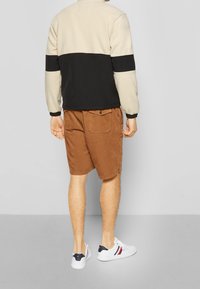 Beige ja musta fleece-takki, rento istuvuus, pitkät hihat. Yhdistetty ruskeisiin ribbineuloshousuihin ja valkoisiin tennareihin, joissa on laivastonsinisiä yksityiskohtia.