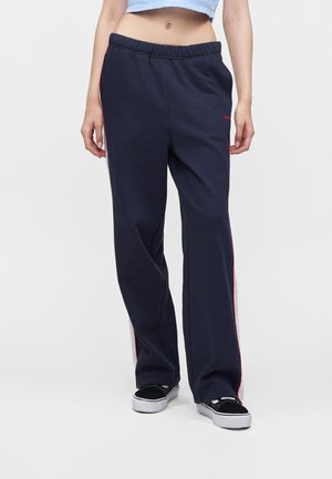 Person iført navyblå løstsiddende joggingbukser med hvide og røde sidelstriber og sorte og hvide sneakers, stående mod en ensfarvet baggrund.