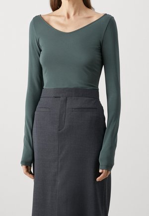 Vrouw draagt een donkergroen shirt met lange mouwen en een brede halslijn, ingestopt in een hooggetailleerde donkergrijze rok met zakken met openingen.