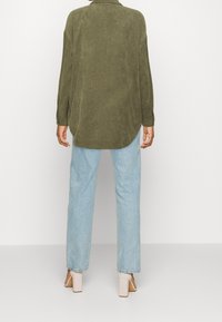 Camisa de pana verde oliva con corte relajado, mangas largas y dobladillo curvado, combinada con unos jeans de pierna recta de color azul claro y tacones gruesos.