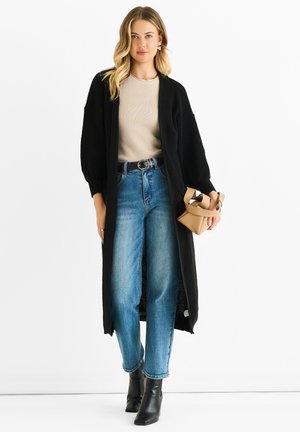 Cardigan nero in maglia sopra una maglietta beige, abbinato a jeans blu a vita alta e stivali neri alla caviglia. Si tiene in mano una piccola borsa tan.