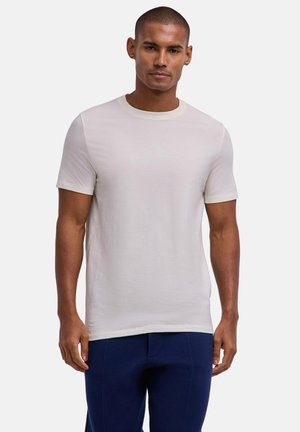 Man met kort haar, gekleed in een eenvoudig wit T-shirt met korte mouwen en donkerblauwe broek, staand tegen een lichtgrijze achtergrond.