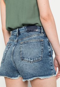 Pantalones cortos de mezclilla en azul claro con un dobladillo deshilachado, que cuentan con bolsillos traseros y una etiqueta de marca negra en la cinturilla. Tejido texturizado.