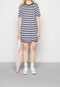 Raitapuku tummansinisen ja valkoisen puuvillan yhdistelmässä, rento malli, lyhyet hihat, helma polvien yläpuolella. Vaaleanpunainen liitospussi yksinkertaisella muotoilulla, yhdistettynä valkoisiin tennareihin.