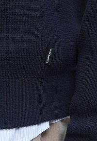 Tamsiai mėlynas megztinis su raštuotu dizainu, su gipiūrinėmis rankovėmis ir apačia. Ant jo matomas juodas audinio etiketė su "JACK & JONES" logotipu.