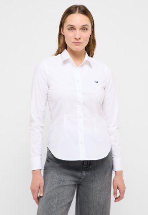 Frau trägt ein eng anliegendes weißes Hemd mit Knöpfen, mit einem kleinen dunklen Logo auf der Brust und graue hoch taillierte Jeans vor einem schlichten Hintergrund.