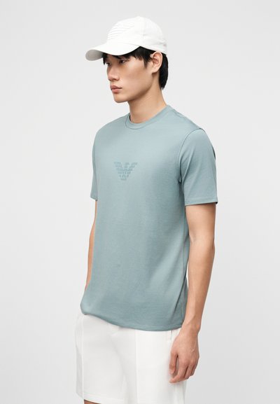 Νεαρός άνδρας που φοράει ανοιχτό μπλε t-shirt Armani με λογότυπο, λευκά σορτς και λευκό καπέλο του μπέιζμπολ, στέκεται μπροστά σε φόντο με απλό χρώμα.