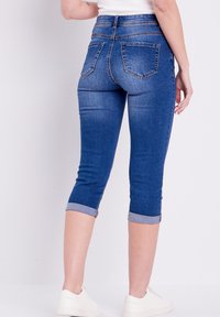 Capris de mezclilla con una silueta ajustada, color azul oscuro, con puños enrollados y dos bolsillos traseros. La tela parece suave con un ligero estiramiento.