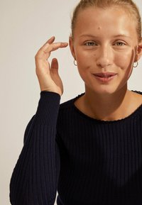 Pull en maille côtelée marine avec un col bateau et des manches ajustées. Gros plan sur un visage avec un maquillage naturel et des boucles d'oreilles créoles, la main près des cheveux.