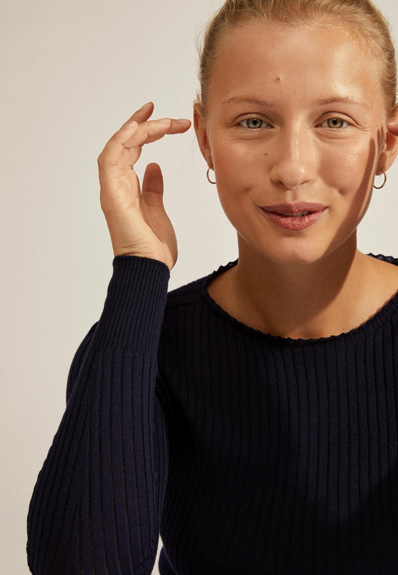 Pull en maille côtelée marine avec un col bateau et des manches ajustées. Gros plan sur un visage avec un maquillage naturel et des boucles d'oreilles créoles, la main près des cheveux.