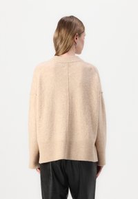 Beige ullgenser med en avslappet passform, ribbede mansjetter og kant. Myk tekstur med skuldresseam som strekker seg inn i ermene. Enkelt design.