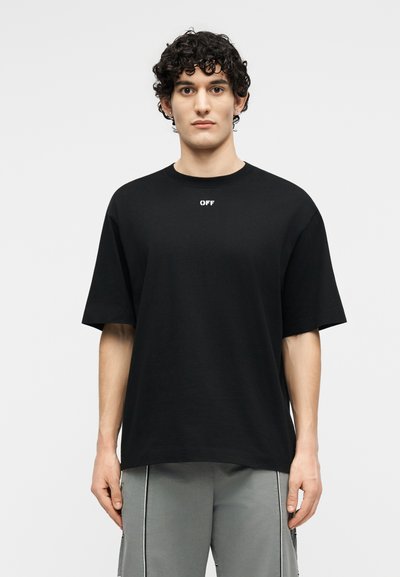 Νεαρός άνδρας με σγουρά μαλλιά που φοράει ένα μαύρο φαρδύ T-shirt με το κείμενο "OFF" κοντά στη γραμμή του λαιμού και γκρι παντελόνι με κάθετες ρίγες.