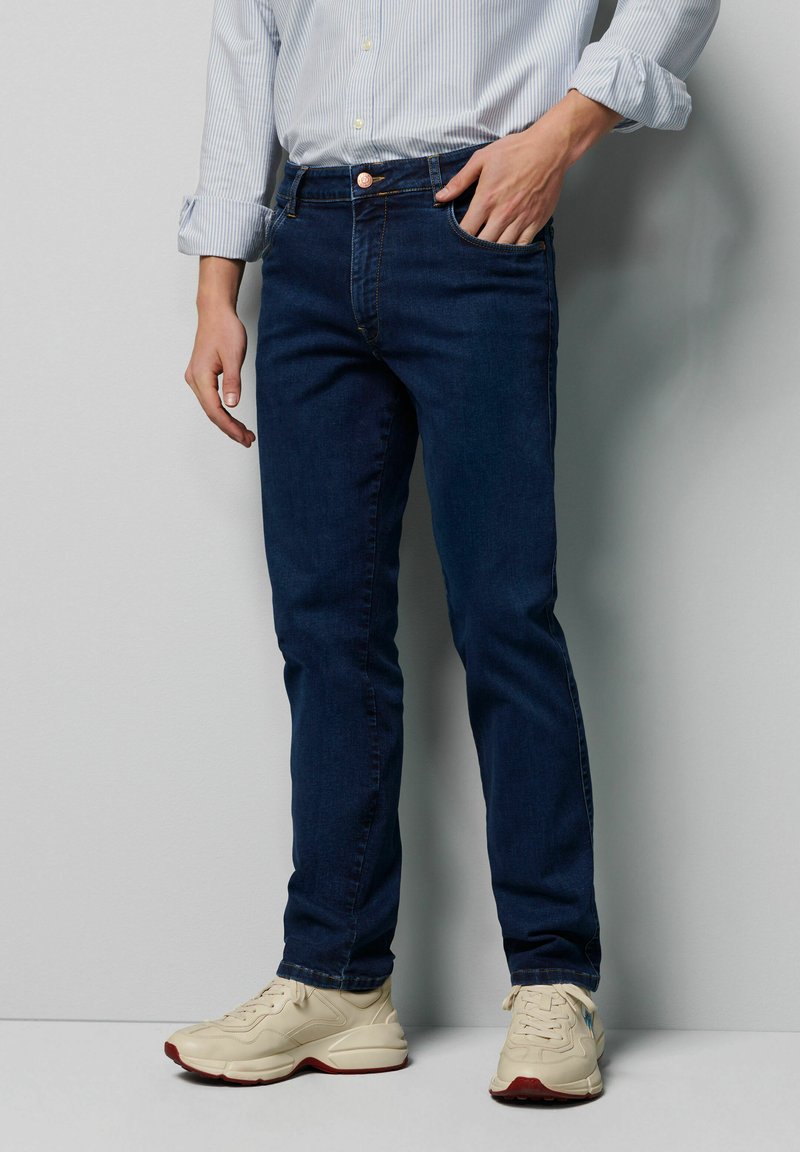 Donkerblauwe denim jeans met een slim fit, vijf zakken en koperen knoopdetails, gecombineerd met crèmekleurige sneakers.