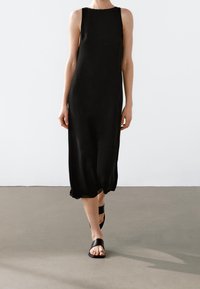 Robe midi en soie noire avec un décolleté rond et sans manches, présentant une silhouette fluide et un détail subtil à l'ourlet. Associée à des sandales noires.
