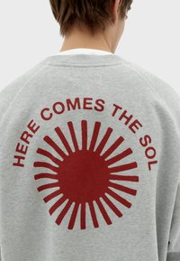 Sudadera gris con un gráfico redondo en la parte posterior que presenta un diseño de sol rojo y un texto que dice "AQUÍ VIENE EL SOL". Textura de tela suave.