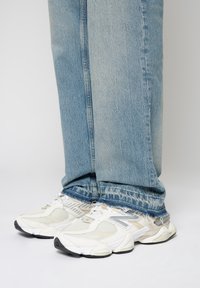 Personne portant un jean bleu clair à ourlet effiloché et des baskets blanches à semelles épaisses avec des accents gris et beige sur un fond blanc.