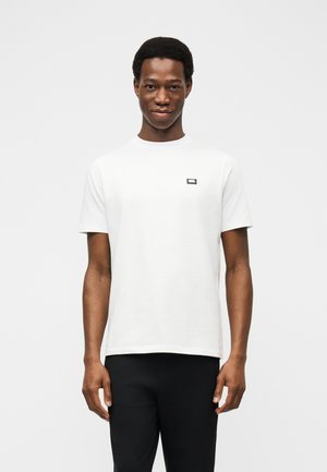 Bărbat stând cu fața înainte purtând un tricou alb simplu cu mâneci scurte și pantaloni negri, pe un fundal alb.