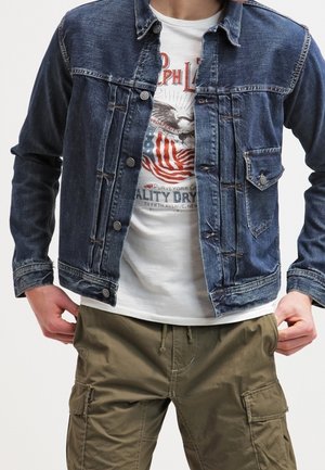 Mężczyzna w niebieskiej dżinsowej kurtce na białym graficznym t-shircie i oliwkowych spodniach cargo, stojący z rękami blisko kieszeni.