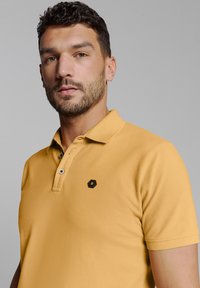 Gele polo met een klassieke kraag, een knoopsluiting met drie knopen en een zwart hexagonaal logoaccent op de borst. Soepele katoenen stof.