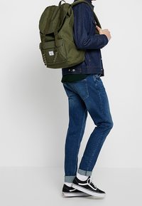 Zielony plecak z tkaniny z dwoma górnymi paskami, przednią kieszenią i ściągaczem. Nosi się go z ciemnoniebieskimi dżinsami i czarnymi sneakerami.