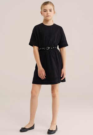 Junges Mädchen steht, trägt ein schwarzes kurzärmeliges Kleid mit einem mit Nieten besetzten Gürtel und schwarze Ballerinas vor einem schlichten hellen Hintergrund.