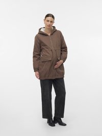 Moteris, apsirengusi rudą gobtuvu striukę, juodas plačias kelnes ir juodus aukštakulnius batus, švelniai laiko savo nėščia pilvą, stovinti ant balto fono.