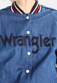 Džínová bunda s velkým vyšitým logem "Wrangler". Tmavě modrá barva, zapínání na cvoky a pruhovaný žebrovaný límec s červenými a bílými akcenty.