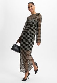 Schwarzes und goldenes Netz-Zweiteiler-Kleid mit langen Ärmeln, Wellenmuster, Seitenschlitzen, kombiniert mit schwarzen High Heels und einer kleinen Handtasche.